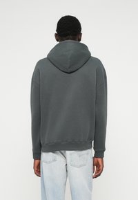 Szary sweter z kapturem o luźnym kroju, z prążkowanymi mankietami i dołem. Materiał wygląda na miękki i teksturowany, noszony z jasnoniebieskimi dżinsami.