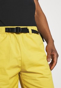 Žluté šortky z texturované látky, s elastickým pasem a černým pásem, s černým tričkem, které je částečně viditelné nahoře.