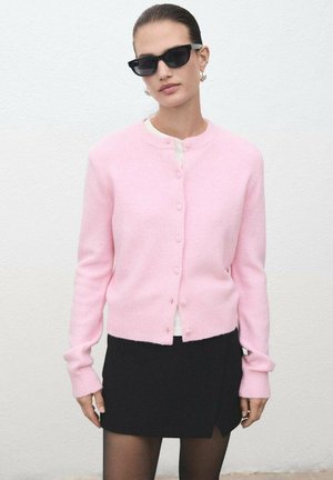 Cárdigan rosa claro hecho de tela suave, con escote redondo, cierre de botones y mangas largas, combinado con una falda negra y gafas de sol.