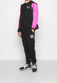 Svart träningsdräkt med en rosa jacka med lång ärm, med en grafisk logotyp, strukturerade svarta byxor och svart-vita sneakers.