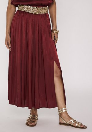 Femme portant une jupe plissée bordeaux avec une fente latérale, une ceinture tissée, des bracelets dorés et des sandales métalliques à brides.