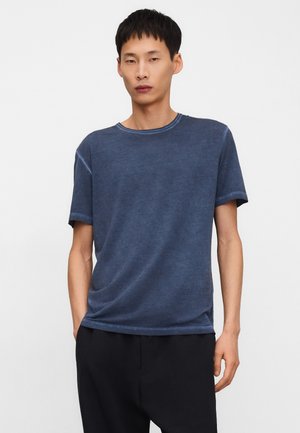 Jonge man met kort zwart haar, gekleed in een lichtbehouden blauw t-shirt met korte mouwen en een zwarte broek, staand tegen een effen witte achtergrond.