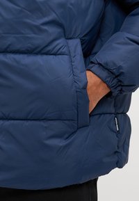 Doudoune bleu marine avec des sections matelassées, des poignets élastiques et une poche latérale visible. Le matériau semble lisse et isolé.