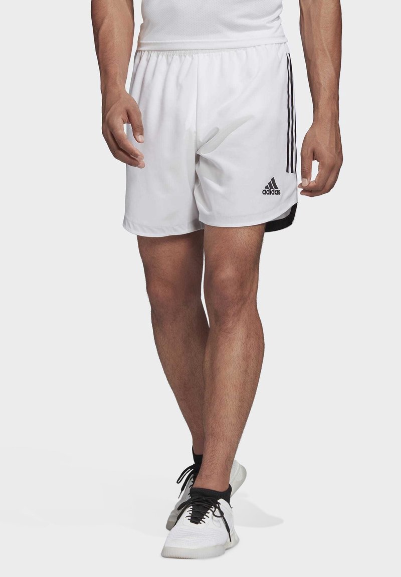 Homme portant un short de sport blanc Adidas avec des bandes noires et des baskets blanches marchant vers l'avant sur un fond uni.