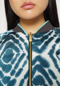 Bomber bunda s tyrkysovým a bílým vzorem, zlatým zipem, šedým límcem a tyrkysovými akcenty podél výstřihu. Tkanina působí hladce a má texturu.