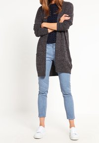 Mörkgrå stickad lång cardigan över en marinblå prickig skjorta, kombinerad med ljusblå ankellånga jeans och vita sneakers.