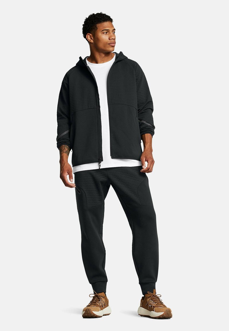 Schwarze Zip-Hoodie mit strukturierten Einsätzen und passenden Joggerhosen; darunter ein weißes T-Shirt und braune Sportschuhe.