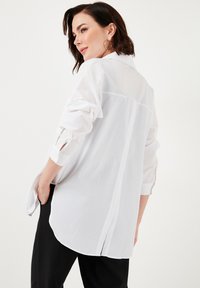 White long-sleeve shirt with a back button closure, soft fabric, curved hem, and rolled sleeves. Worn with black pants.