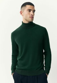Pulover verde cu guler înalt, din material moale, având un design ajustat și manșete și tiv elastic, purtat lejer peste pantaloni bleumarin.