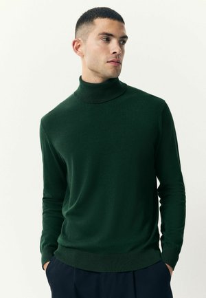 Suéter verde de cuello alto, hecho de material suave, con un diseño ajustado y puños y dobladillo acanalados, llevado de manera informal sobre pantalones azul marino.