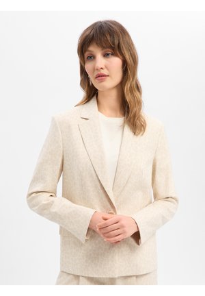 Vrouw met schouderlang golvend haar die een lichtbeige gebloemde blazer en bijpassende broek draagt, staand met handen gevouwen.
