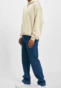 Sudadera color crema con un bolsillo frontal, combinada con jeans azules y zapatillas blancas. La sudadera cuenta con cordones y puños acanalados.