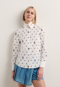 Hvit langermet skjorte med små fargerike tegneserie mønstre, med spiss krage og broderte røde detaljer. Kombinert med denimbukser med folder.