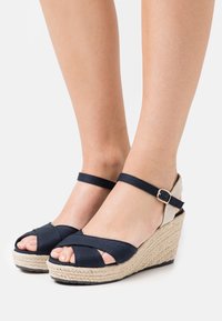 Sandales compensées bleu marine avec design à lanières croisées, semelle enveloppée de jute, et bride de cheville réglable avec une boucle dorée en accent.