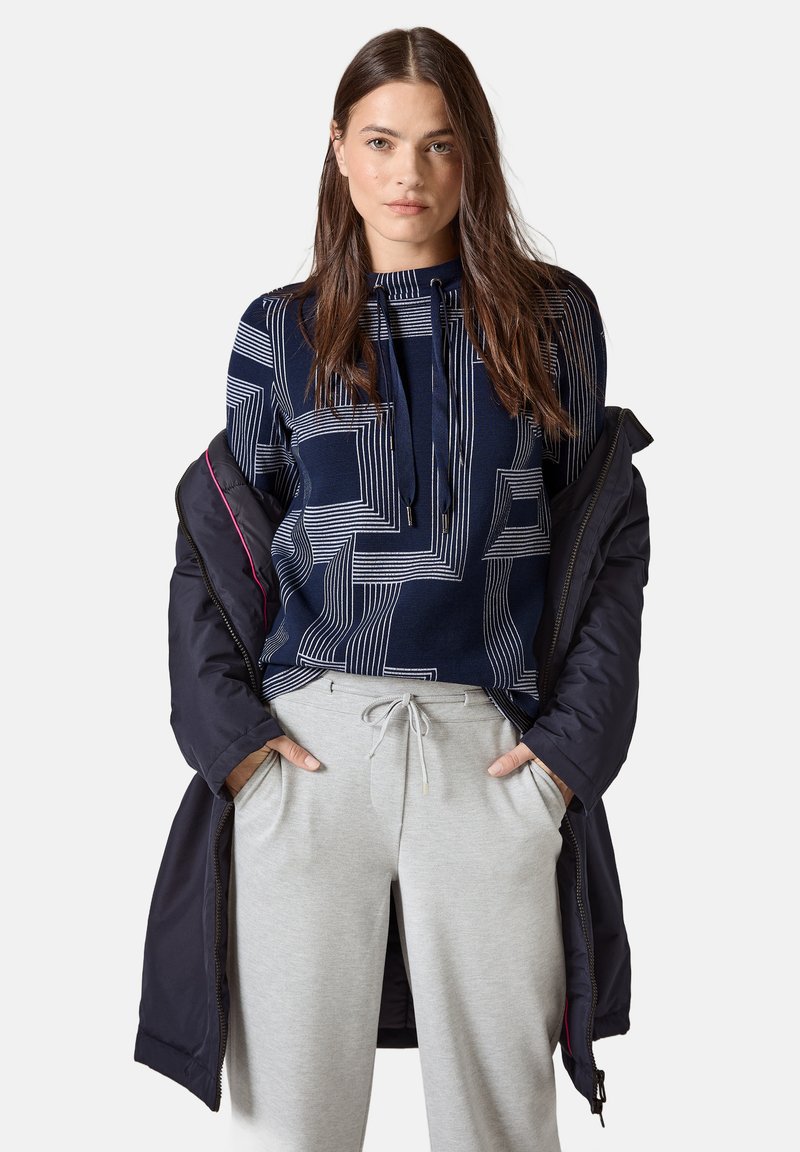 Navyfarbener Kapuzenpullover mit weißen geometrischen Linien, kombiniert mit hellgrauen Jogginghosen. Die äußere Bekleidung ist in Dunkelblau mit pinken Akzenten.