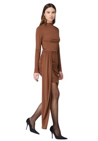 Braunes langärmliges Kleid mit gerafftem Design und asymmetrischem Saum, aus weichem Material. Kombiniert mit schwarzen High-Heels und durchsichtigen Strumpfhosen.