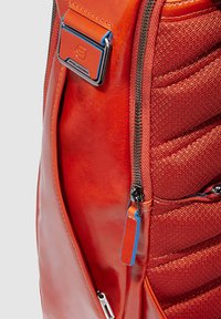 Mochila de cuero roja con paneles de tela texturizada, herrajes metálicos, cremalleras visibles y etiqueta azul de acento.