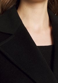 Cappotto di lana nero con colletto strutturato, caratterizzato da una texture liscia e un design minimale; indossato sopra un top nero a coste.