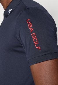 Polo azul-marinho feito de um tecido liso, com um bordado vermelho "USA GOLF" no ombro direito e um design de colarinho contrastante.
