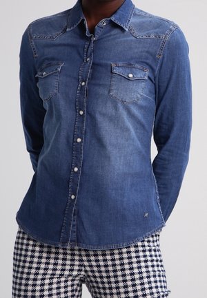 Person trägt ein tailliertes blaues Denim-Hemd mit Knöpfen und Brusttaschen sowie kariere schwarze und weiße Hose.