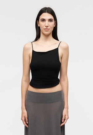 Nő hosszú sötét hajjal, fekete ujjatlan felsőt és szürke magasderekú szoknyát visel, és egyszínű világos háttér előtt áll.