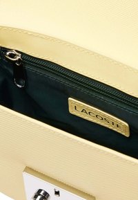Die Innenseite der Lacoste-Handtasche zeigt ein blassgelbes strukturiertes Futter, eine dunkelgrüne Reißverschlusstasche und ein goldenes Etikett mit dem Markennamen.
