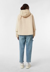 Cappa in misto lana beige con cappuccio trapuntato, abbinata a pantaloni in denim azzurro chiaro. Il design oversize presenta maniche ampie e una vestibilità rilassata.