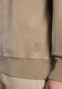 Sweatshirt beige avec des poignets et un ourlet côtelés, arborant un logo en relief sur le bas à gauche. Tissu doux avec une texture lisse.
