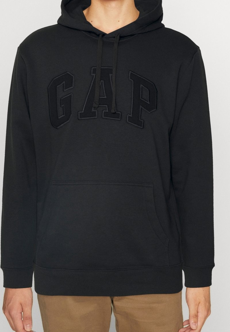 Svart hoodie med framficka och svart broderad "GAP"-text, bärs med tanfärgade byxor, visad från halsen till höften.
