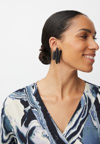 Orecchini in polimero nero dalla forma ovale allungata, caratterizzati da tre elementi sovrapposti. La persona che li indossa ha i capelli all'indietro.