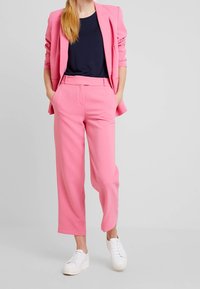 Blazer rosa ajustado y pantalones a juego, ambos de tela suave. Combinado con una blusa azul marino y zapatillas blancas. Corte entallado y recto.