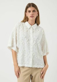 Blusa bianca a maniche corte con colletto, caratterizzata da ricami floreali, dettagli a fessura e una vestibilità ampia. Tessuto texturizzato con accenti morbidi.