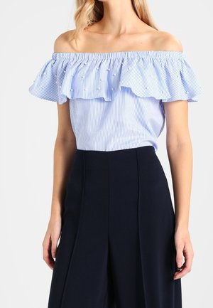Frau trägt ein blau-weiß gestreiftes Off-Shoulder-Top mit gerafftem Ausschnitt und schwarze, weit geschnittene, hoch taillierte Hose.