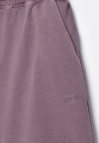 Gros plan sur un pantalon de survêtement mauve montrant une poche latérale et un petit logo de marque brodé près de la poche.