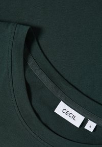 Camiseta verde oscuro con escote redondo. Tejido suave con una textura lisa. La etiqueta dice "CECIL" con la talla "S" visible.