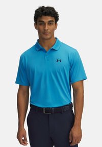 Světle modré polo tričko s límečkem a krátkými rukávy, s černým logem Under Armour na levé straně hrudi. Nosené s tmavými kalhotami.