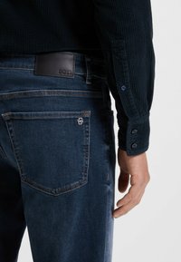 Donkerblauwe denim jeans met een leren "BOSS" label. Heeft een vijf-pocket ontwerp en zichtbare stiksels. Draag ze met een donker, gestructureerd knoopshirt.