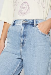 Jeans bleu clair taille haute avec bouton et fermeture éclair à l'avant, portés avec un t-shirt beige pâle de la marque Lee Company, main droite dans la poche.