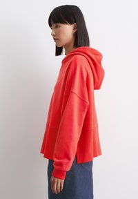 Rode hoodie met een ontspannen pasvorm, geribbelde manchetten en zijsplitten, gecombineerd met een spijkerrok. De stof lijkt zacht en licht gestructureerd.