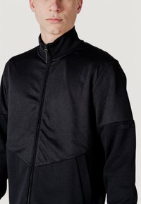 Giacca nera con zip, caratterizzata da una parte superiore in mesh testurizzato e una parte inferiore liscia, con un accento di marca sulla manica.