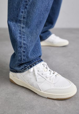 Adidași din piele albă cu accente texturate, talpă din cauciuc gri deschis, design cu șireturi și purtați cu blugi albaștri.