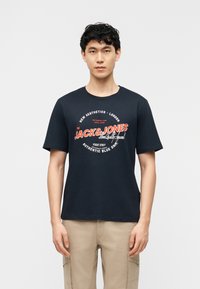 Νέος άνδρας που φοράει μαύρο μπλουζάκι Jack & Jones με στάμπα και μπεζ παντελόνι cargo, στέκεται μπροστά σε μια απλή λευκή επιφάνεια.