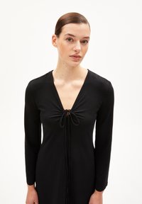 Schwarz langärmliges Kleid mit tiefem V-Ausschnitt, das eine geraffte Vorderseite und Bindebänder aufweist. Der Stoff wirkt glatt und weich. Minimalistisches Design.