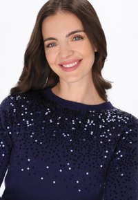 Tengerés kék pulóver bordás textúrával, az alsó részén fokozatos mintázatban elhelyezett flitteres díszítésekkel.