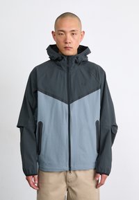 Nike jacket featuring a color-block design in dark gray and light blue, zippered front, adjustable hood, and side pockets.
