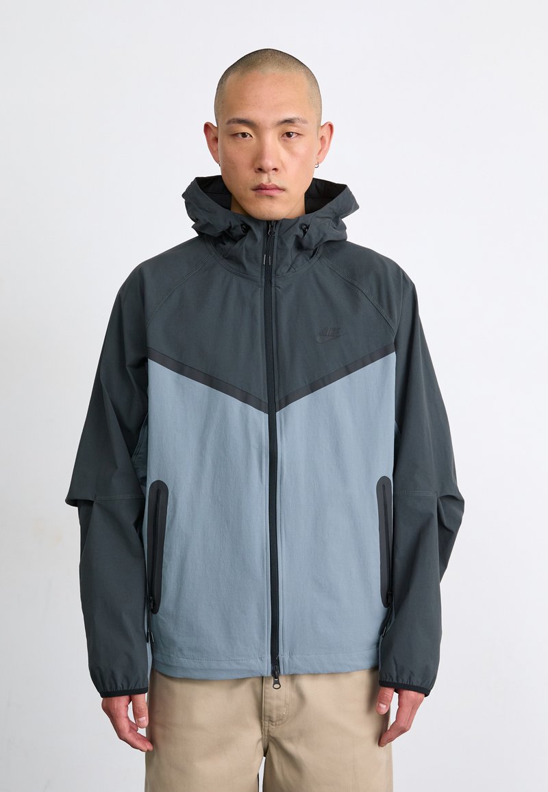 Nike jacket featuring a color-block design in dark gray and light blue, zippered front, adjustable hood, and side pockets.
