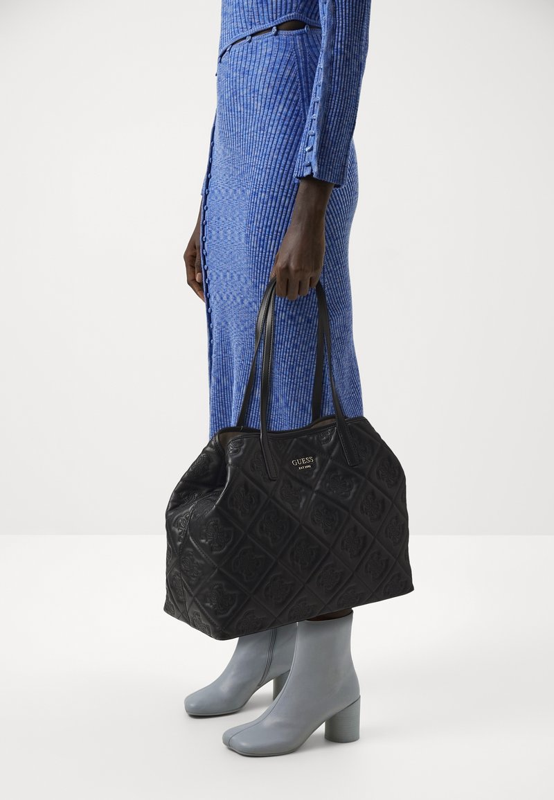 Personne en robe bleu côtelée et bottines grises tenant un grand sac cabas noir Guess avec motif embossé et doubles poignées.