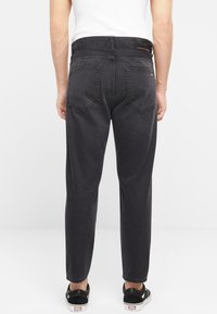 Jeans de mezclilla negra con un corte recto, que presentan dos bolsillos traseros y una etiqueta de marca en la cinturilla, combinados con zapatillas blancas y negras.