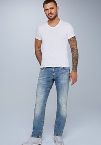 Camiseta blanca de cuello en V, jeans azules desgastados con costuras visibles y zapatillas grises. Modelo mostrado de pie contra un fondo claro.