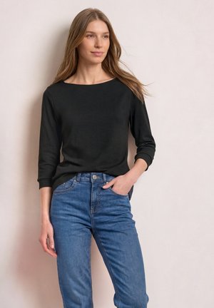 Femme aux longs cheveux portant un haut noir à manches longues et un jean bleu, debout avec une main dans sa poche devant un fond uni.